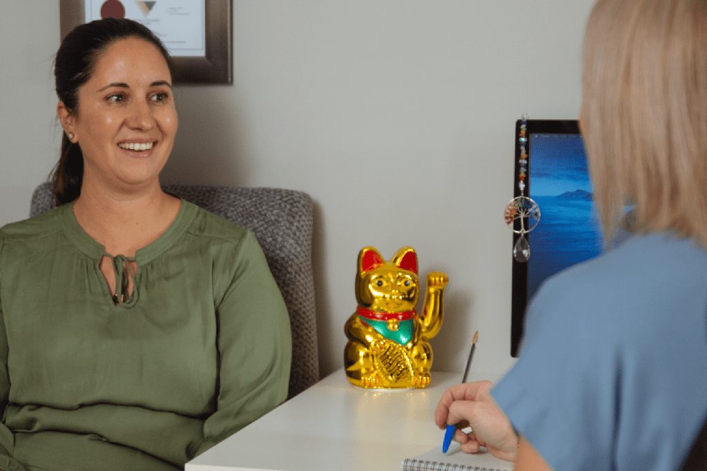 hypnotherapy, macarthur complete health, therapy, hypnosis, Hypnotherapist, Smiling woman in moss-green blouse talking to her therapist, who is seated at her desk and writing down notes